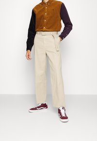 Camisa de pana marrón y negra con botones, pantalones de cintura alta beige y zapatillas burdeos con detalles blancos. Presentación de un conjunto casual.