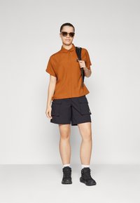 Brown short-sleeve button-up shirt, black cargo shorts with pockets, black boots, sunglasses, standing against a light gray background.