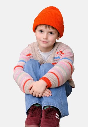 Child sitting cross-legged, wearing an orange knit beanie, multicolored striped sweater, light blue jeans, and burgundy sneakers.