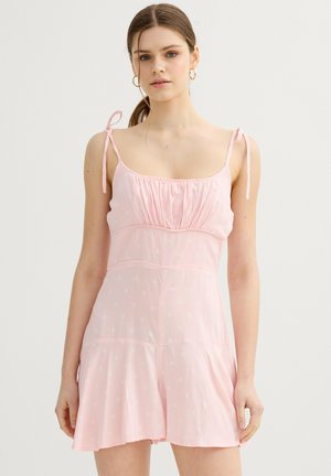 Joven mujer con el pelo recogido, que lleva un vestido sin mangas de color rosa claro con tirantes finos y sutiles patrones florales blancos.