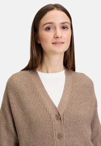Vaffelstickad, beige cardigan med V-ringning, utrustad med två bruna knappar och en avslappnad passform. Texturerad tyg med ribbade accenter.