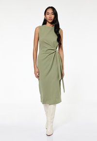 Vestido sin mangas de color verde oliva con un detalle de frente retorcido y un lazo lateral, hecho de una tela suave. Combinado con botas crema de caña alta.