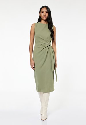 Vestido sin mangas de color verde oliva con un detalle de frente retorcido y un lazo lateral, hecho de una tela suave. Combinado con botas crema de caña alta.