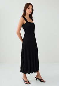 Schwarzes, plissiertes Midikleid mit quadratischem Ausschnitt, breiten Trägern und strukturiertem Stoff. Hat ein enges Oberteil und einen ausgestellten Rock, kombiniert mit schwarzen Sandalen.