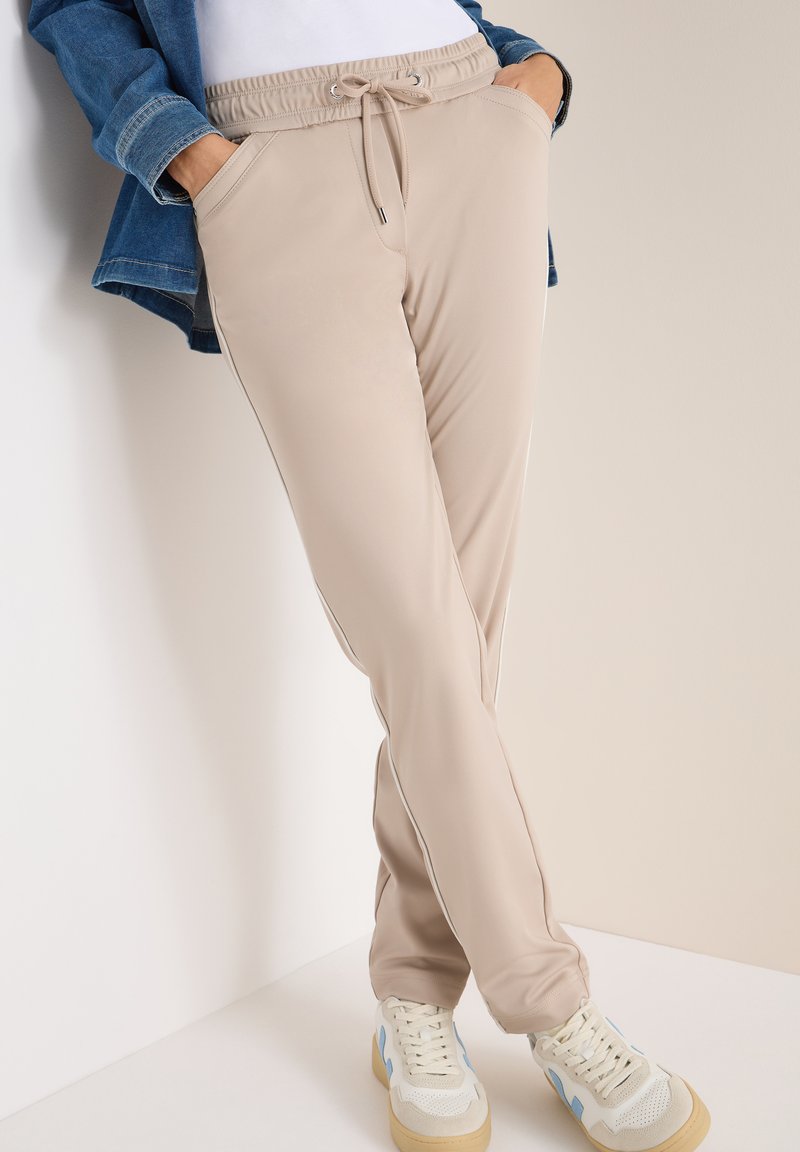 Béžové tepláky s elastickým pasem, stahovací šňůrkou, dvěma bočními kapsami a bílými postranními pruhy; nošeny s bílými teniskami s modrými akcenty.