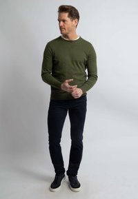 Olijfgroen sweater met een ronde hals, gedragen met donkere jeans en zwarte sneakers. Eenvoudig, aangesloten ontwerp, gladde textuur, geen zichtbare patronen.