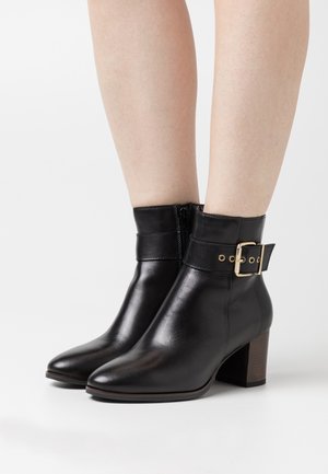 Bottes de cheville en cuir noir avec un accent en boucle dorée, texture lisse, bout pointu et un talon carré de hauteur moyenne. Fermeture éclair sur le côté.