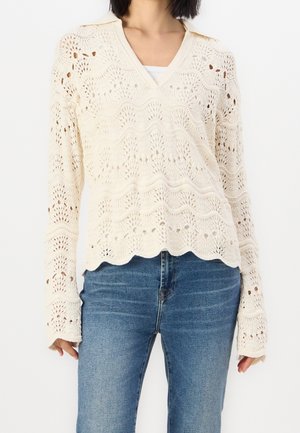 Žena v krémovém, dlouhém svetrů s háčkováním s ozdobným lemem a modré džíny na jednoduchém bílém pozadí.