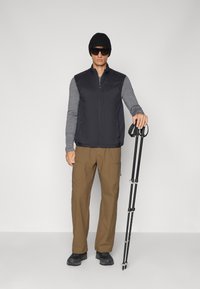 Zwarte geïsoleerde vest over een grijs longsleeve shirt, bruine cargobroek en zwarte laarzen. Houdt een zwarte wandelstok vast, draagt een zonnebril en een beanie.