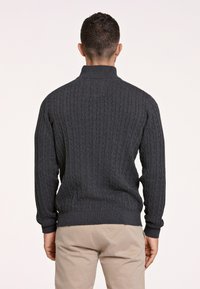 Dunkelgrauer, gestrickter Pullover mit einem strukturierten Zopfmuster, hohem Kragen sowie gerippten Bündchen und Saum, getragen mit hellen Hosen.