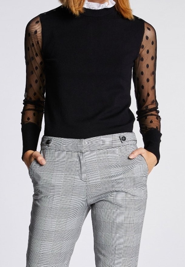 Femme portant un haut noir avec des manches longues transparentes à pois et un pantalon taille haute à carreaux gris, les mains dans les poches.