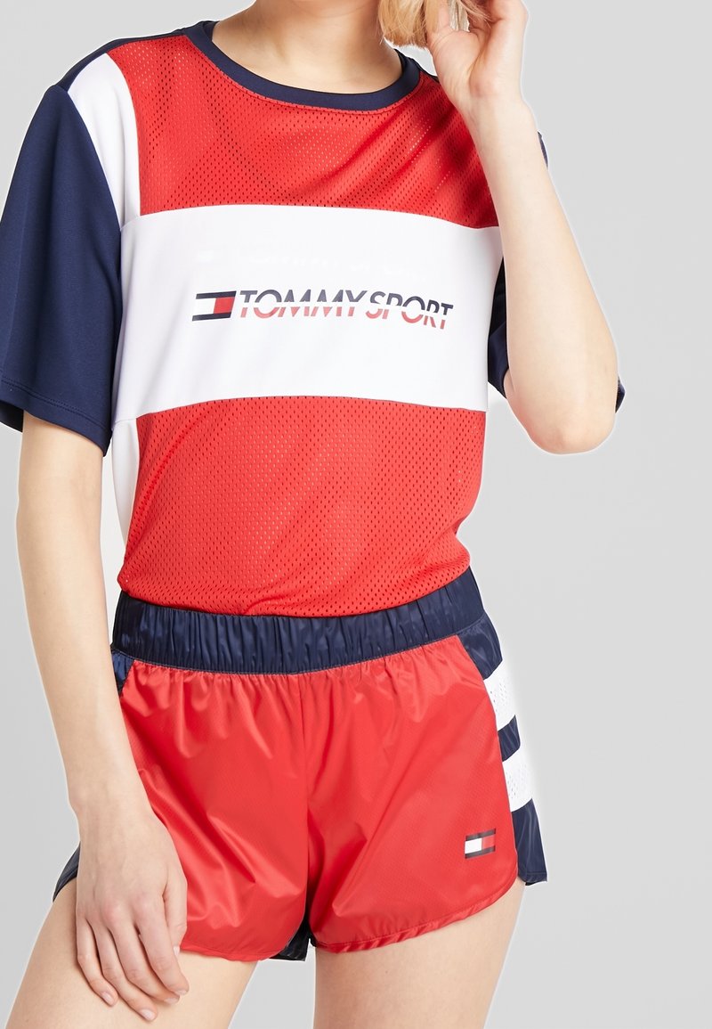 Czerwony i granatowy strój sportowy z siateczkową koszulką z krótkim rękawem w białe paski oraz logo, zestawiony z dopasowanymi czerwonymi szortami z bocznymi paskami.