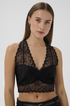 Giovane donna con capelli castani lisci che indossa un top nero in pizzo corto con collo halter e orecchini a cerchio dorati su uno sfondo grigio.