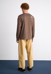 Braunes Langarmshirt mit Rundhalsausschnitt und kleinem Logo am oberen Rücken, kombiniert mit lockeren gelben Hosen und schwarzen Schuhen.