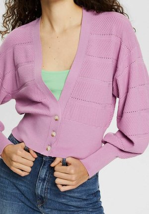 Vrouw draagt een lavendelkleurige, opengewerkte, knoopjes-cardigan over een groen topje met blauwe spijkerbroek, handen rustend nabij de taille.