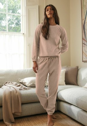 Conjunto de loungewear beige claro y texturizado que incluye una parte superior de manga larga con puños acanalados y pantalones con cintura ajustable, usado en interiores por una modelo.