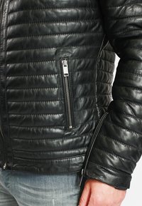 Gros plan d'une veste en cuir noire matelassée avec des surpiqûres horizontales, une poche zippée, et une main reposant près de l'ourlet.