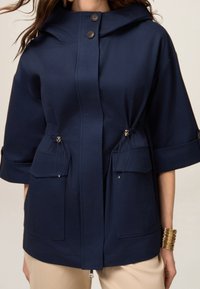 Veste à capuche bleu marine avec une fermeture à boutons sur le devant, deux poches à cordon, des manches courtes et un tissu lisse et texturé.