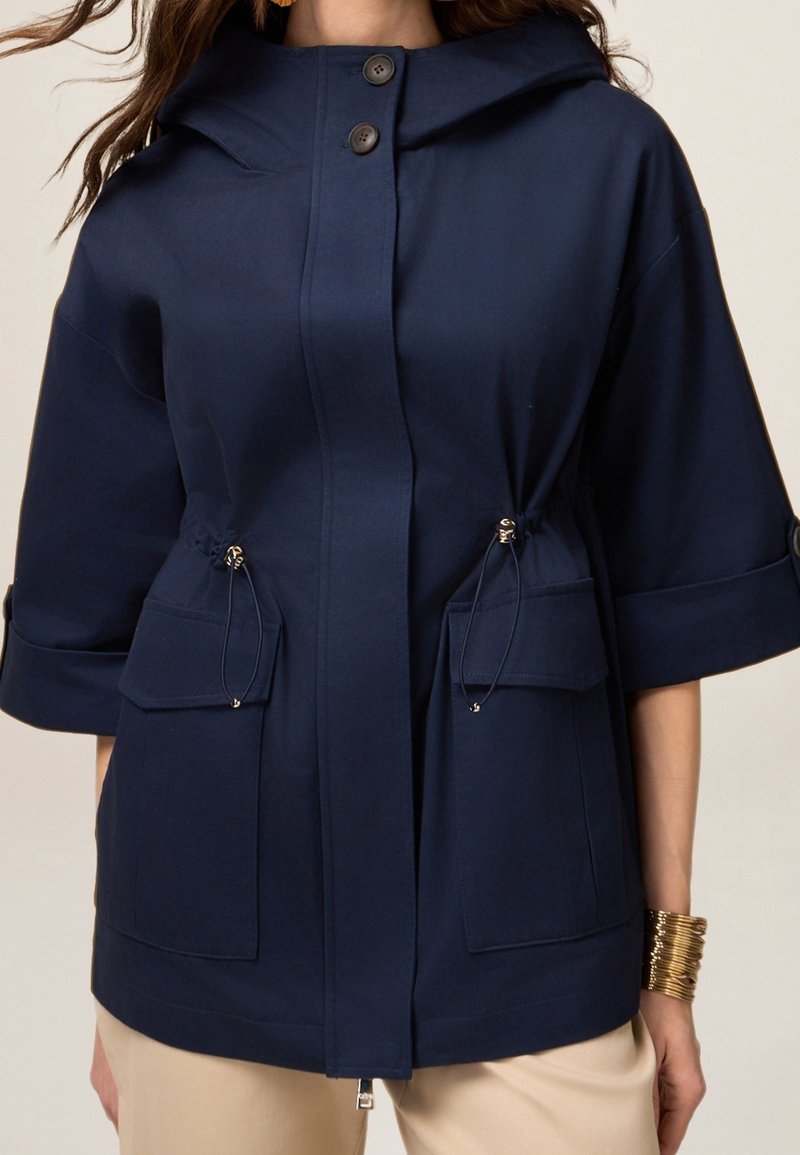 Veste à capuche bleu marine avec une fermeture à boutons sur le devant, deux poches à cordon, des manches courtes et un tissu lisse et texturé.