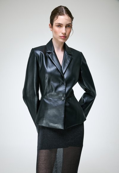 Femme aux cheveux bruns portant un blazer en cuir noir et une jupe noire transparente, debout, les mains dans le dos, sur un fond clair uni.