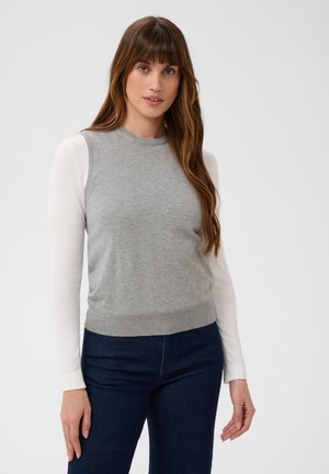 Kvinde med langt brunt hår iført en grå ærmeløs sweater over en hvid langærmet skjorte og mørkeblå jeans, står mod en ensfarvet baggrund.