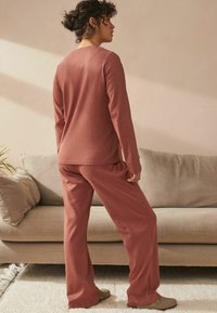 Rosa Loungewear-Set mit langen Ärmeln und elastischem Bund, das eine subtile Textur und eine lockere Passform aufweist, kombiniert mit neutralen Schuhen.