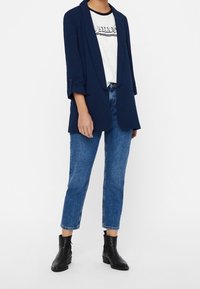 Granatowa marynarka o luźnym kroju, noszona na białej koszulce z nadrukiem, w połączeniu z przyciętymi niebieskimi dżinsami i czarnymi botkami za kostkę.