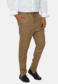 Pantaloni in lino beige con vita elasticizzata, tasche laterali e vestibilità affusolata. Abbinati a scarpe mocassino nere e una camicia bianca con bottoni.