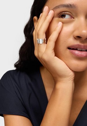 Anillo de plata con un diseño ondulado que alterna secciones pulidas y mate, llevado en el dedo índice sobre una prenda oscura.