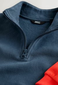 Mørkeblå fleece halv-zip pullover med en struktureret overflade og kontrasterende orange manchetter, forsynet med en sort lynlås med et solidt håndtag.