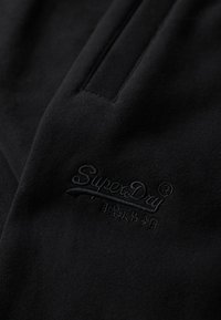 Zwart fleece stof met een gladde textuur beschikt over een subtiele geborduurde "SuperDry"-logo in bijpassend garen. Eenvoudig ontwerp met een ritszak.