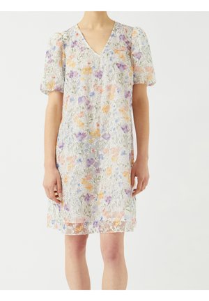 Femme portant une robe blanche à fleurs avec des manches bouffantes et un col en V, ornée de motifs floraux violets, orange et verts sur un tissu transparent.