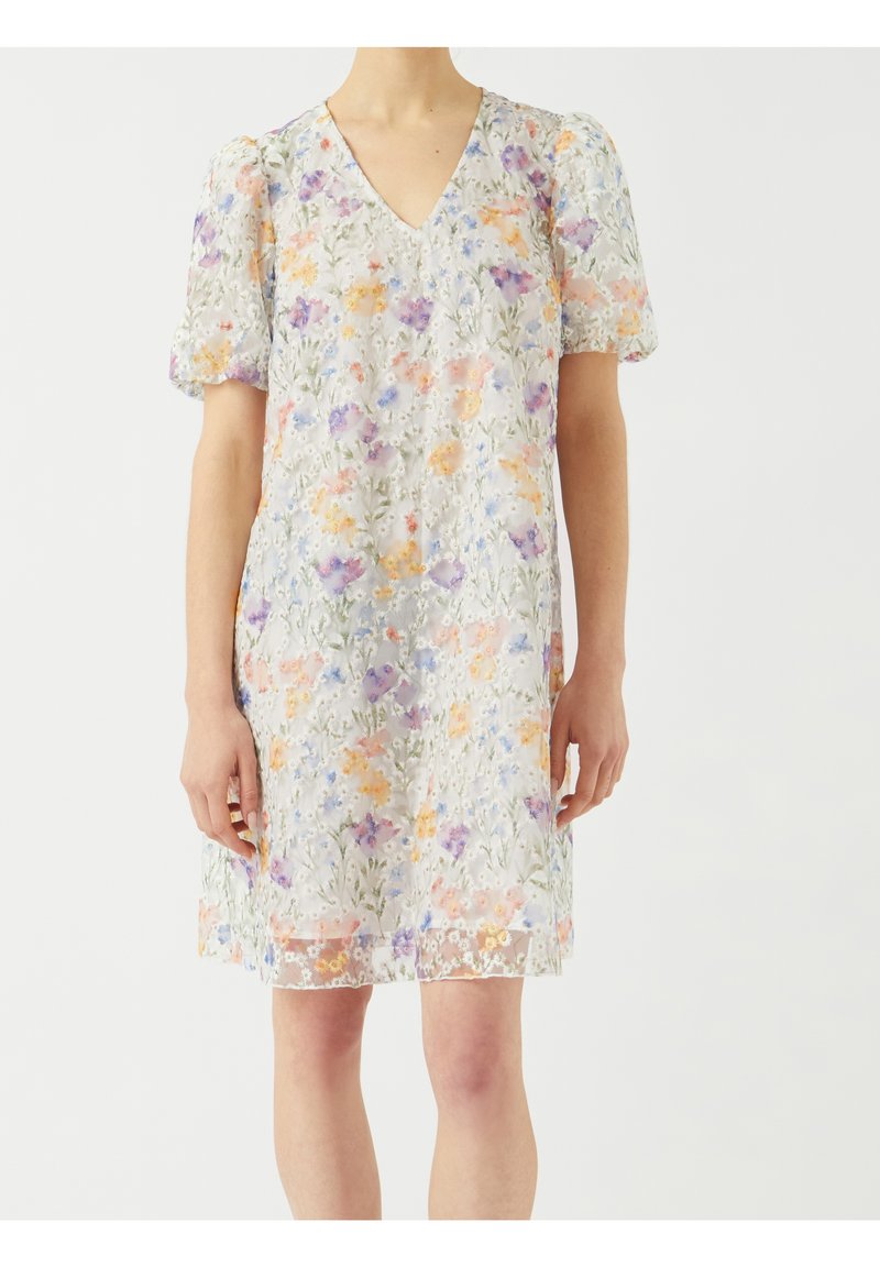 Femme portant une robe blanche à fleurs avec des manches bouffantes et un col en V, ornée de motifs floraux violets, orange et verts sur un tissu transparent.