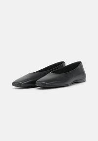 Paire de chaussures plates en cuir noir avec bouts pointus et talons bas sur un fond clair uni.