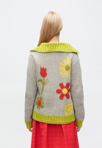 Pulover din lână cu corp gri, guler texturat mare verde și modele florale colorate în roșu, galben și verde. Tiv texturat evidențiat.