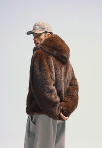 Brun fuskpälsjacka med huva och textured finish, bärs över grå löst sittande byxor, kompletterad med en mönstrad keps.