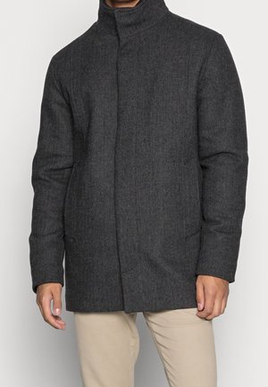 Mężczyzna ubrany w ciemnoszary wełniany płaszcz jodełkowy z wysokim kołnierzem i beżowe spodnie na jednolitym tle.