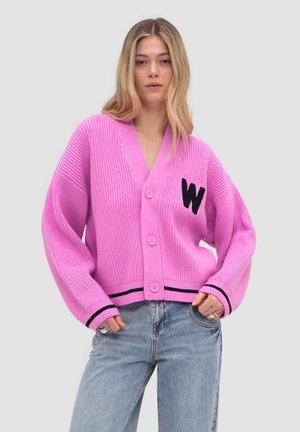 Femme portant un cardigan rose vif boutonné avec un "W" noir sur la poitrine, associé à un jean bleu clair sur un fond uni.
