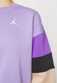 Sudadera morada con bloques de color superpuestos: morado claro, morado oscuro, negro y beige. Incluye un logo blanco y tiene una textura suave.