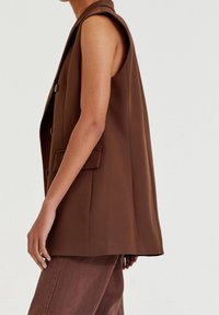 Brown sleeveless blazer with a notched collar, two front pockets, and a relaxed fit, made of smooth fabric. Paired with matching brown pants.