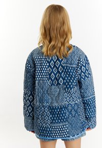 Blå patchworkjakke med forskellige geometriske mønstre, detaljeret syning og tekstureret stof. Designet inkluderer en blanding af lys blå og mørk blå nuancer.