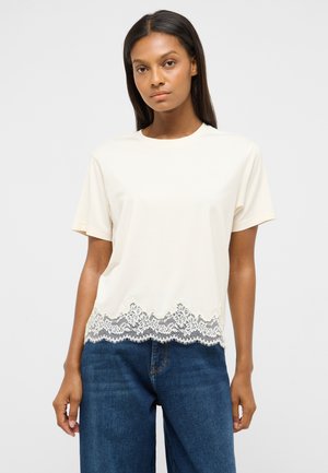 Woman wearing a cream short-sleeve top with lace trim at the hem and blue jeans, standing against a plain white background.