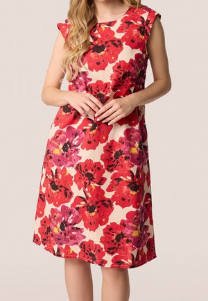 Mouwloze jurk tot op de knie met grote bloemenprint in rood en paars op een lichtbeige achtergrond, gedragen door een vrouw met gevouwen handen.