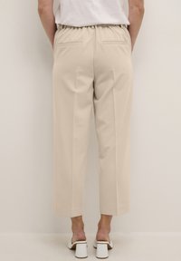Pantalons beiges crop avec une ceinture élastique, deux poches arrière et une texture lisse. Portés avec des sandales à talons carrés blanches.