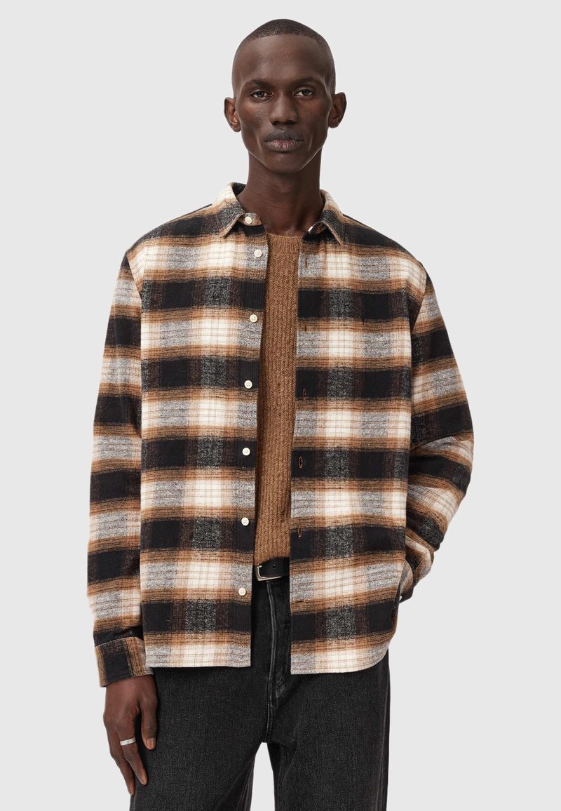 Flanellskjorta med ett svart, brunt och vitt rutmönster, knappstängning framtill och en avslappnad passform, bärs över en brun stickad tröja.