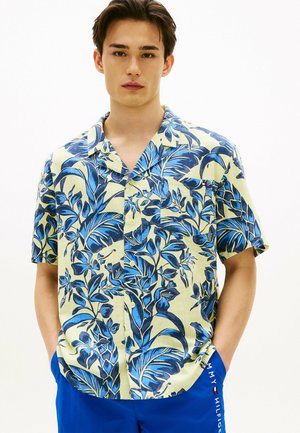 Junger Mann trägt ein kurzärmeliges Hemd mit blauem tropischem Blattmuster und blaue Shorts mit Tommy Hilfiger-Logo.