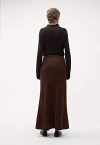 Maglione marrone a coste con maniche lunghe, abbinato a una lunga gonna fluida di color marrone scuro. L'outfit presenta tessuti morbidi e un design semplice.