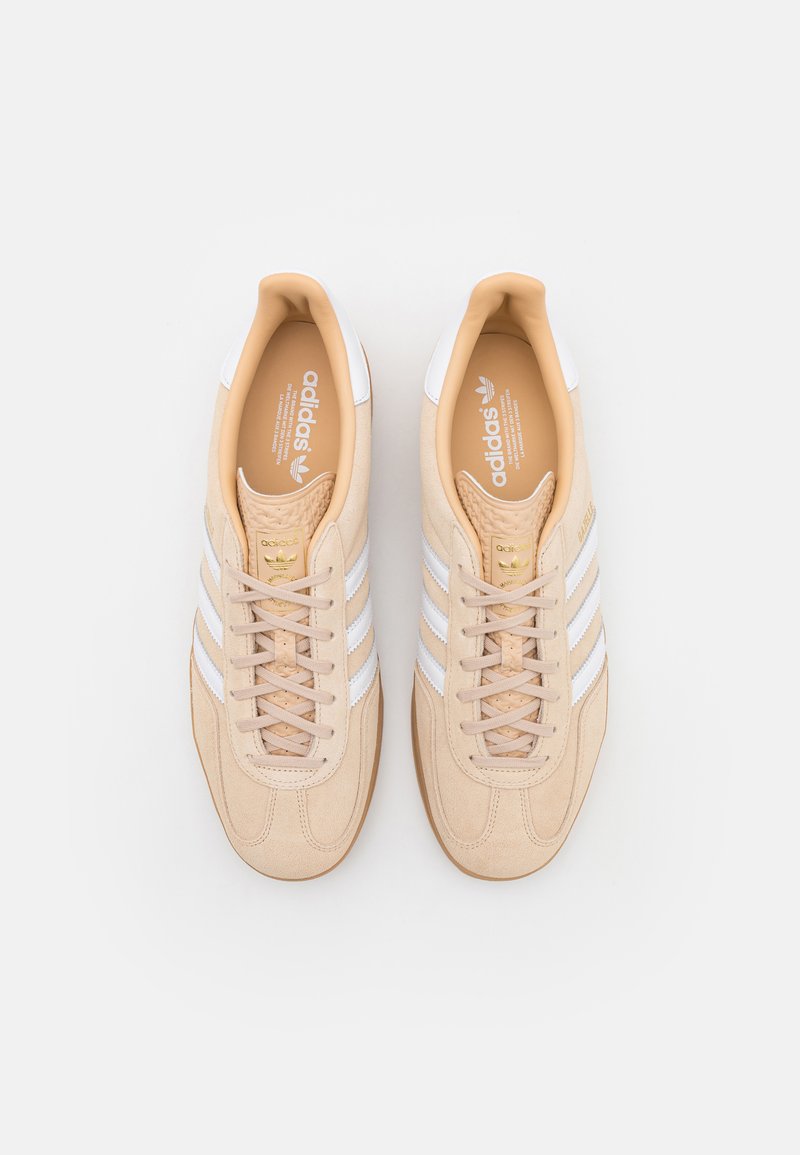 Zapatillas de ante beige con detalles en cuero blanco, que presentan tres franjas blancas, cierre de cordones y una puntera texturizada. Detalle del logo en dorado en el interior.