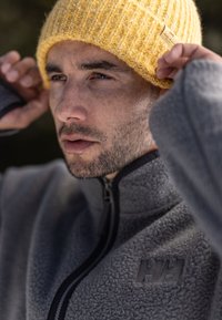 Grijze fleece jas met een zwarte rits en een reliëflogo, gecombineerd met een gestructureerde gele wollen muts. Close-up van een gezicht met stoppels.