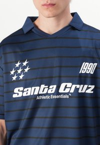 Marine stripete polo t-skjorte med krage. Har hvit tekst "Santa Cruz" og "1990," med fem hvite stjerner på venstre side. Mykt stoff.